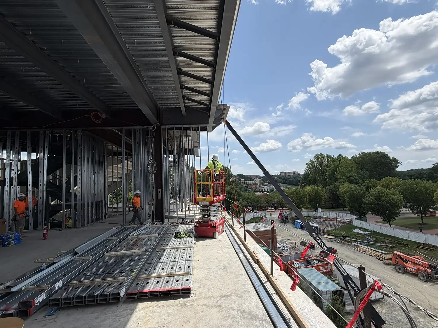Construction workers working on a building structure with steel framing, using a crane and lifted platform, against a backdrop of trees and a partly cloudy sky.