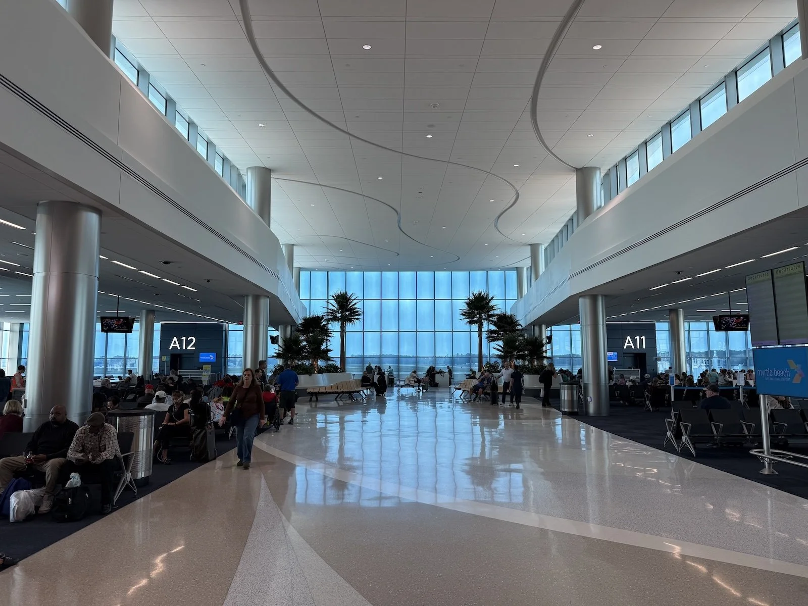 Interior of an airport terminal with high ceilings, large windows, seating areas, and people waiting or walking around. Signs display gate numbers A12 and A11.