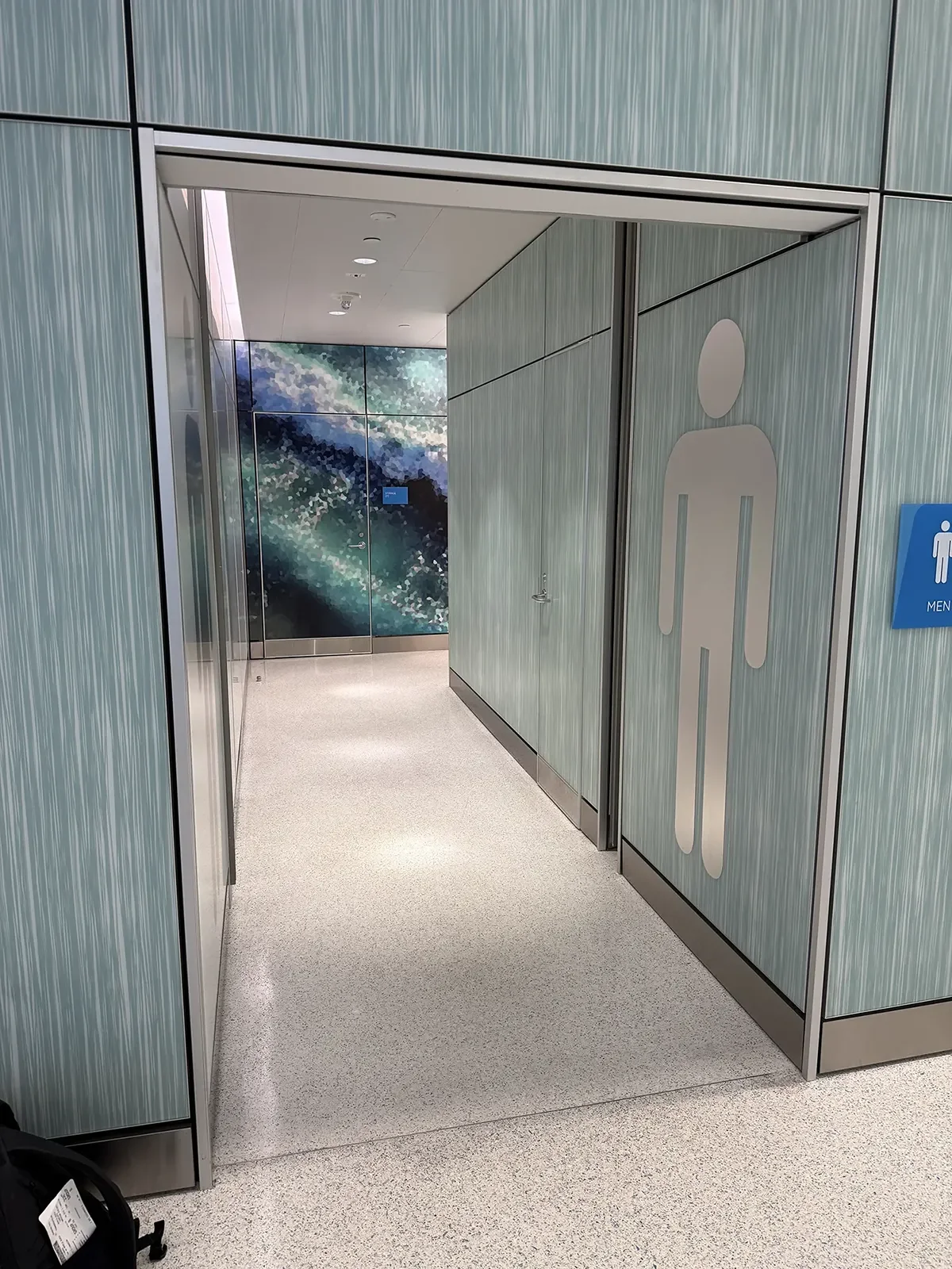 A hallway in a modern building with textured green walls, a light-colored speckled floor, and a large mural of a misty landscape on the back wall. There is a sign indicating men's restroom.