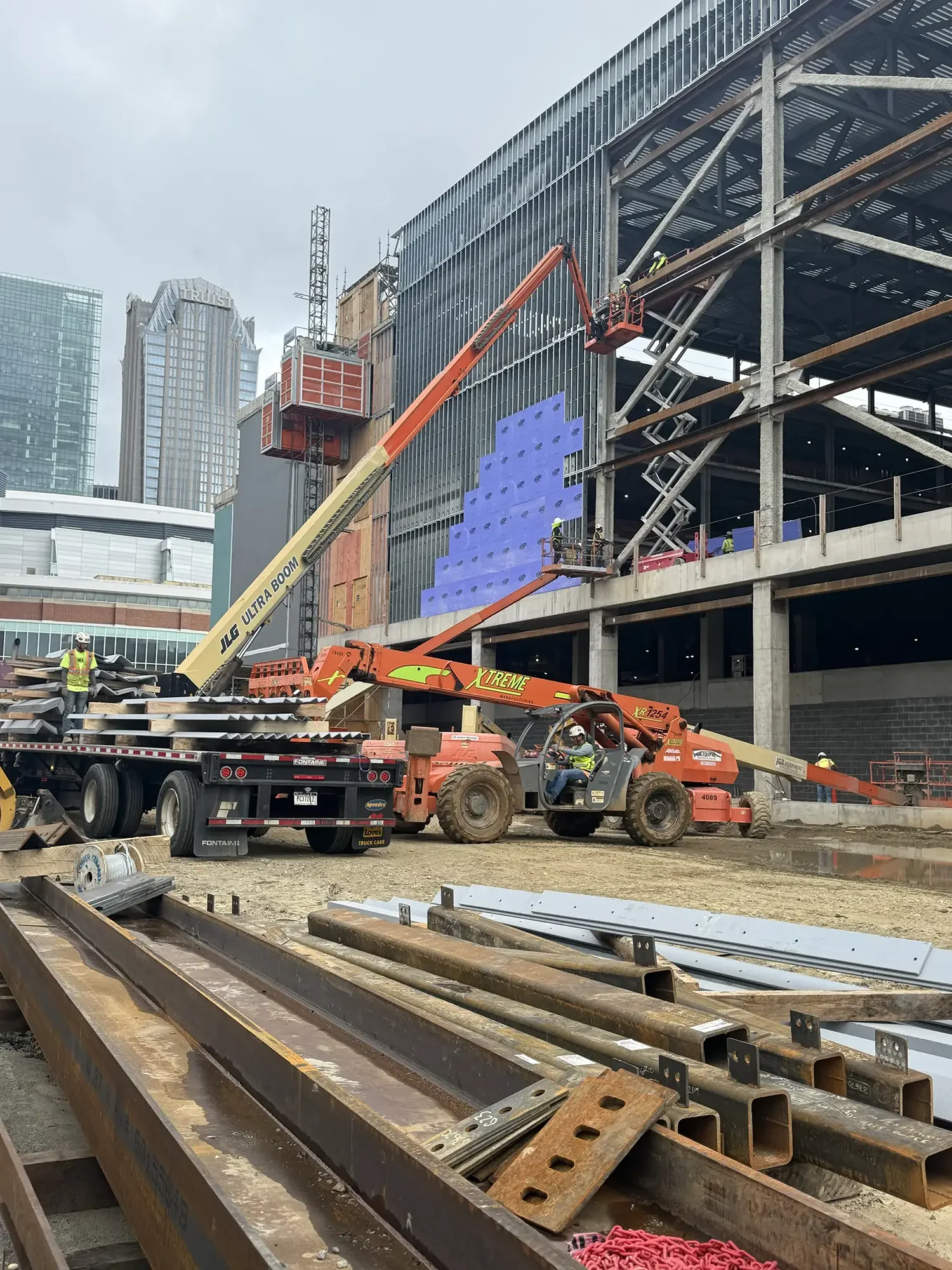 Construction site with workers on a steel framework building, using a crane and telehandler for assembly, and metal beams in the foreground.