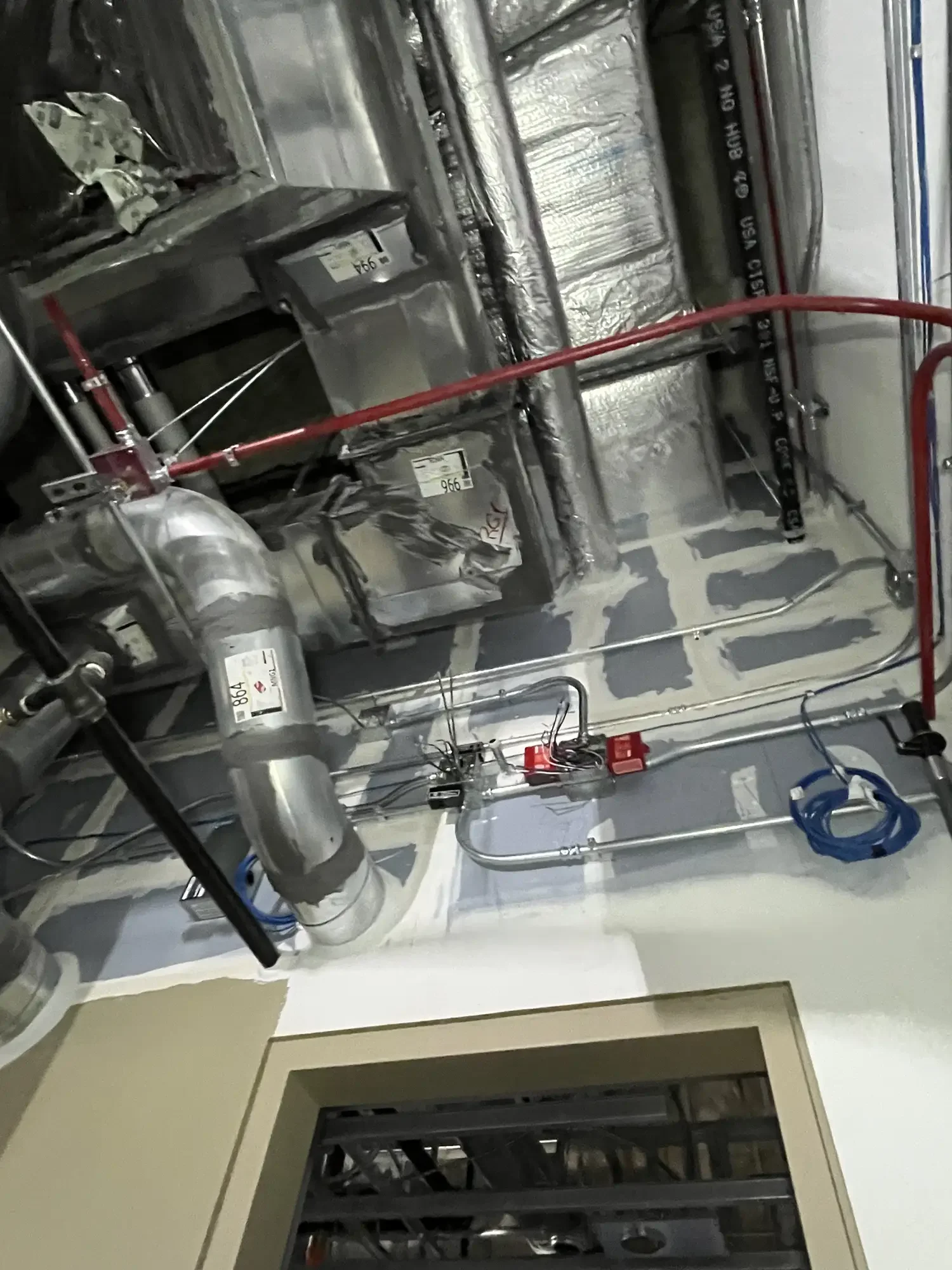 This image shows the interior of an unfinished or maintenance area, with visible ductwork, wiring, and piping. There is a cut-out in the ceiling with some insulation and metal framing, and a doorway below leading into another part of the building.
