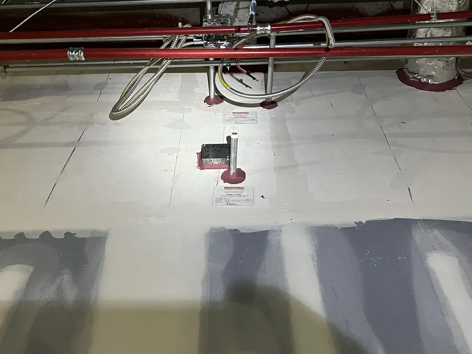 Construction site ceiling with exposed pipes and wiring, insulation seen in the background, and warning labels on the ceiling