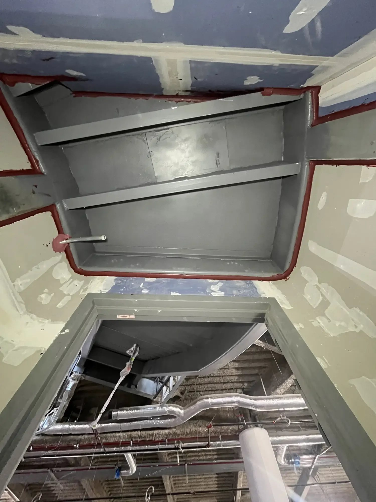 Construction site showing an elevator shaft with unfinished drywall and insulation, exposed pipes, and electrical wiring.