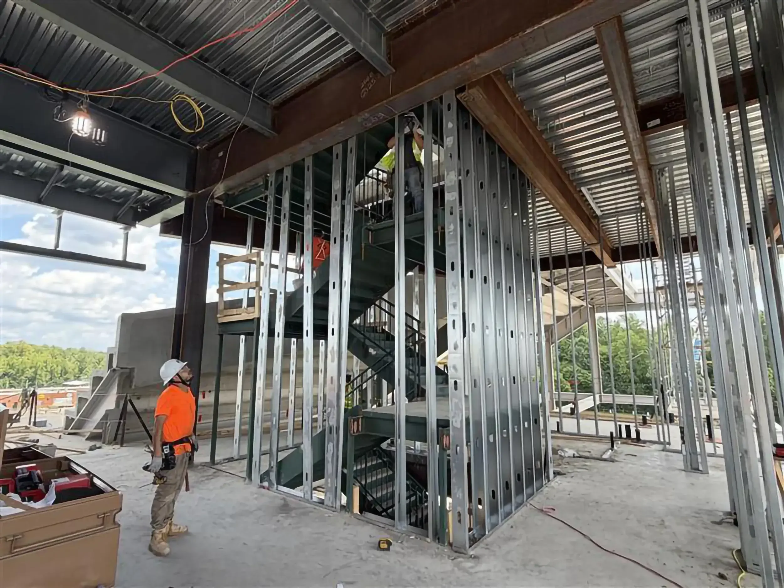 Construction workers framing a multi-story building with metal studs, exposed wiring, and a spiral staircase under a corrugated metal roof