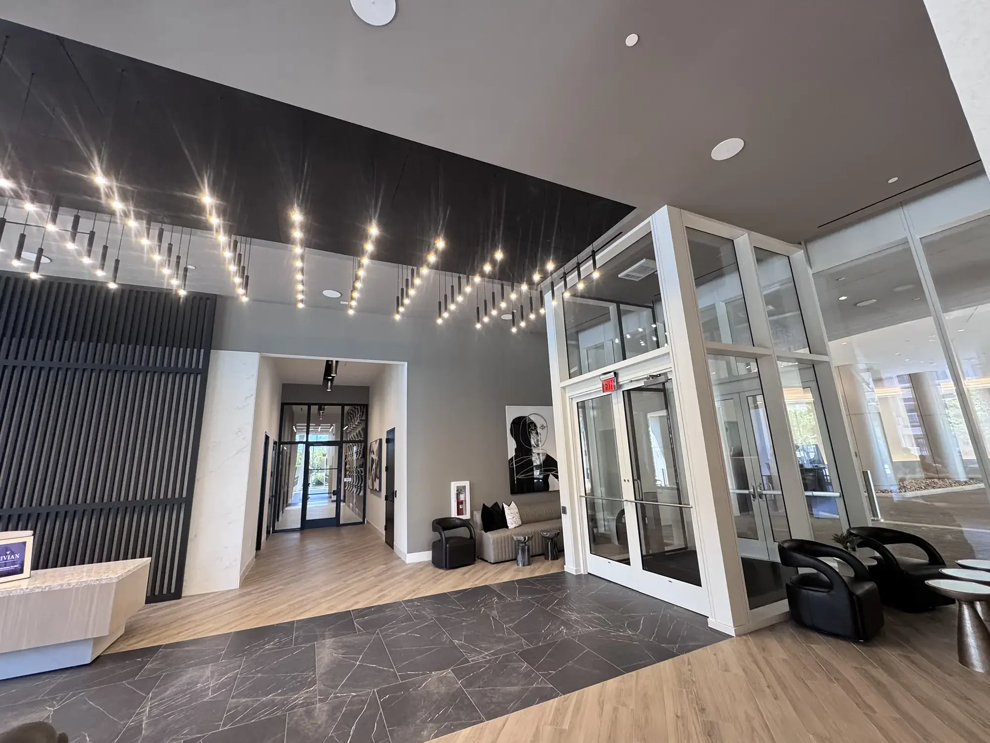 Interior of a modern lobby with a high ceiling, decorative lighting, glass doors, black chairs, and a wooden and marble floor.