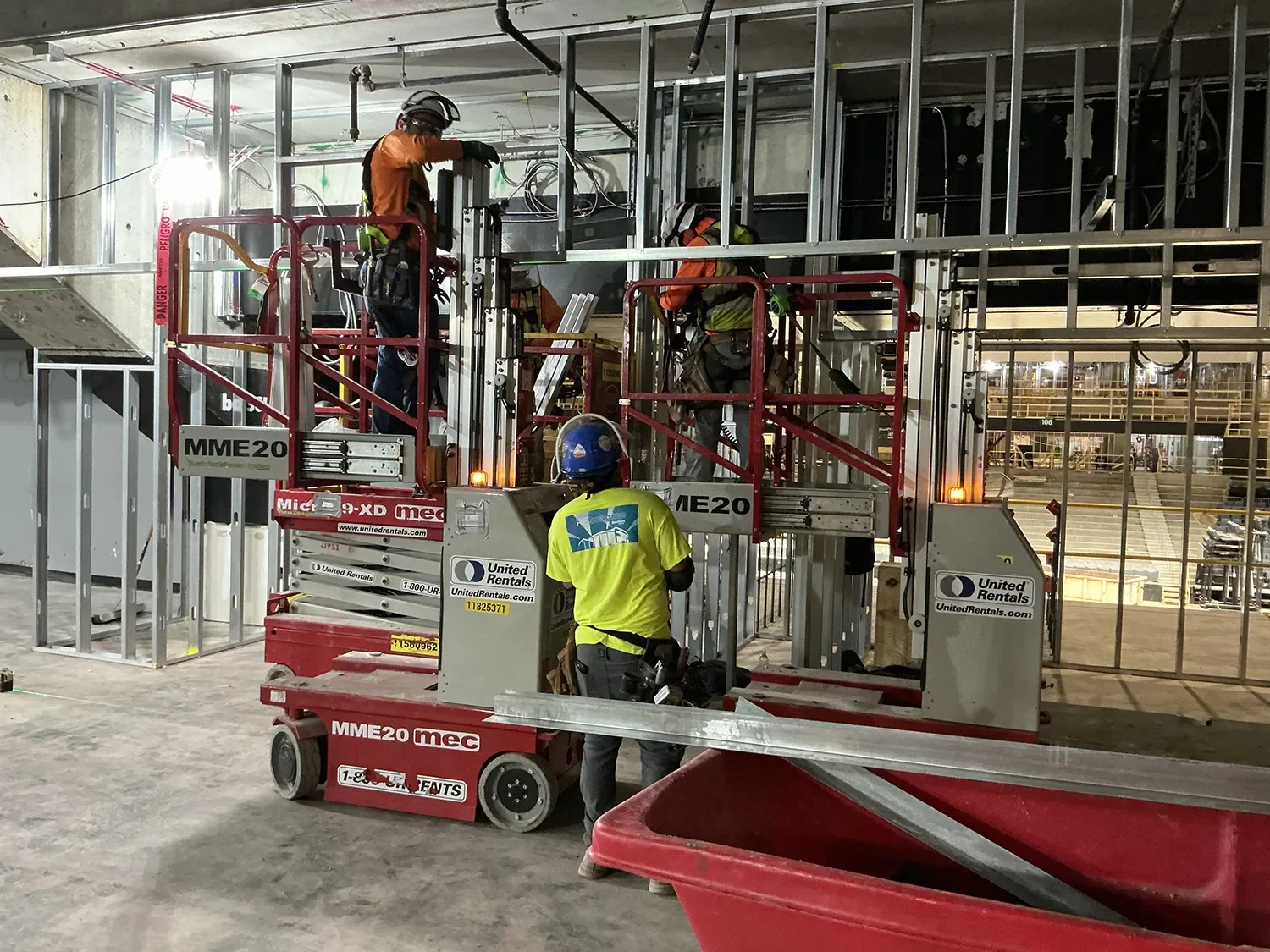 Construction workers in safety gear working on scaffolding at an indoor construction site, with metal framework and equipment around.