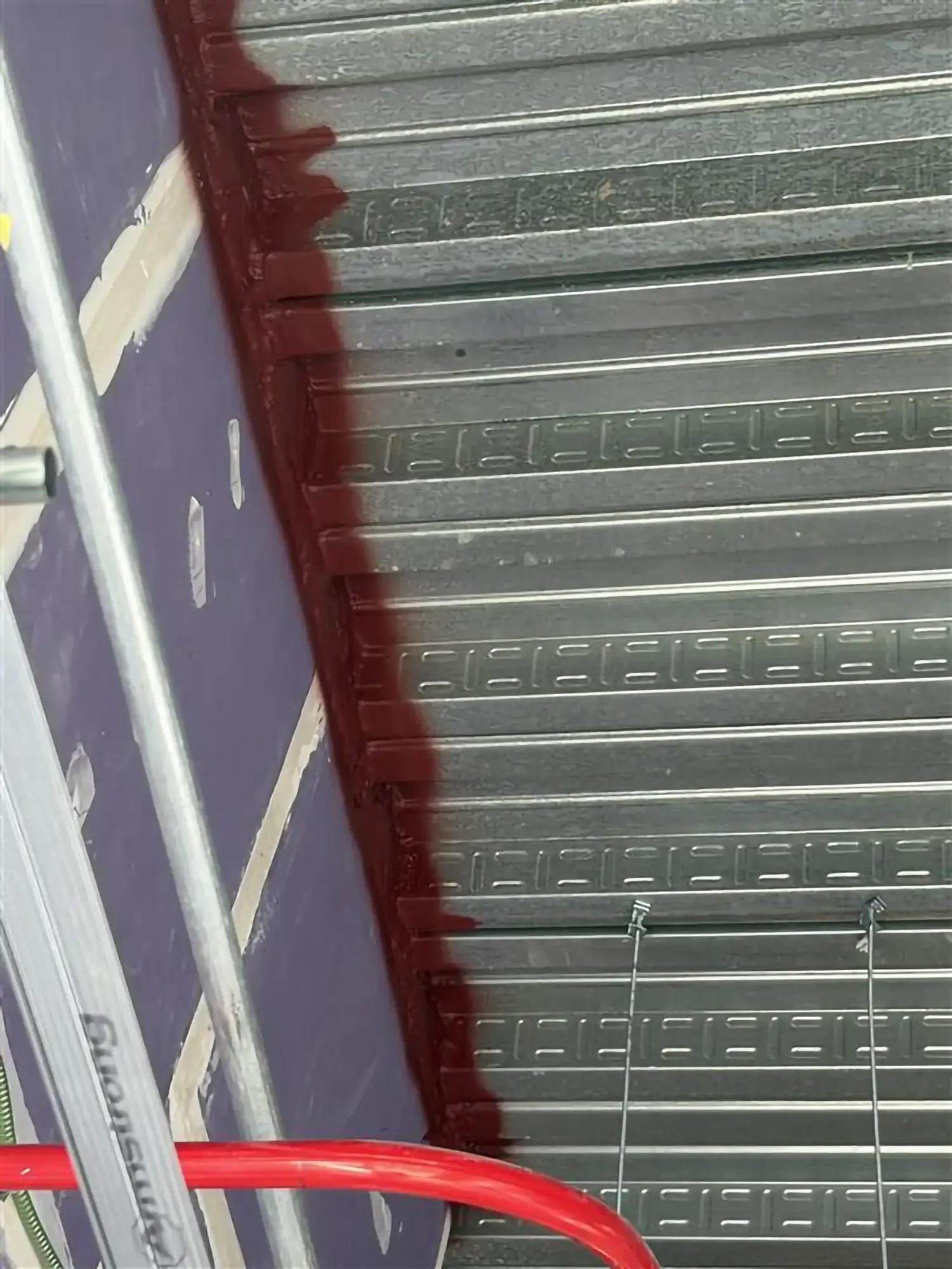 Close-up of a flexible corrugated metal panel with a dark red painted edge and a section of a nearby ledge or ladder.