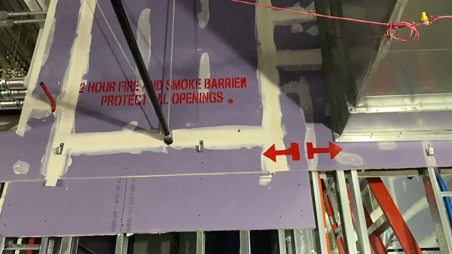 Construction site with purple fire-resistant board marked with red text '2 HOUR FIRE AND SMOKE BARRIER PROTECT ALL OPENINGS' and orange arrows, metal studs, and structural framing.