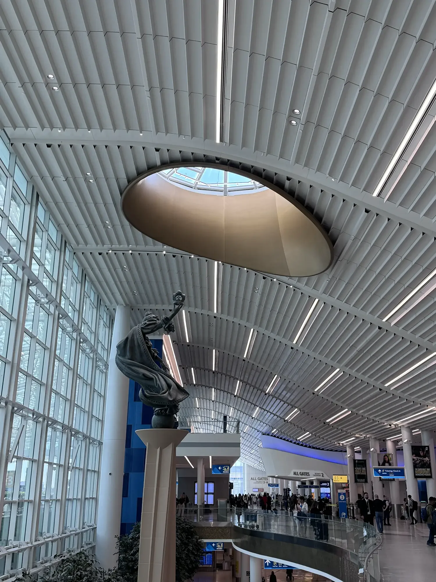 Interior of an airport terminal with a high ceiling, large windows, an oval skylight, and a statue of a seated figure.