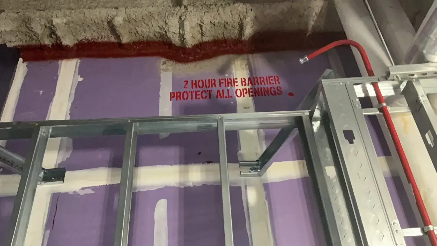 Construction site with metal framing, exposed wall, and a sign that reads '2 HOUR FIRE BARRIER PROTECT ALL OPENINGS'.