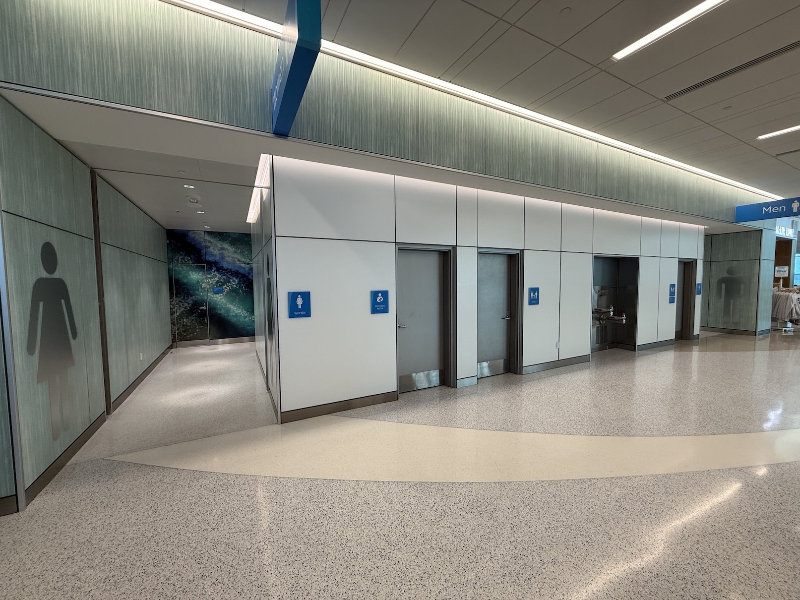 Public restroom area with entrances for women and men, and a gender-neutral option, in an airport or similar facility. Water fountain nearby, and signage indicating restroom facilities.