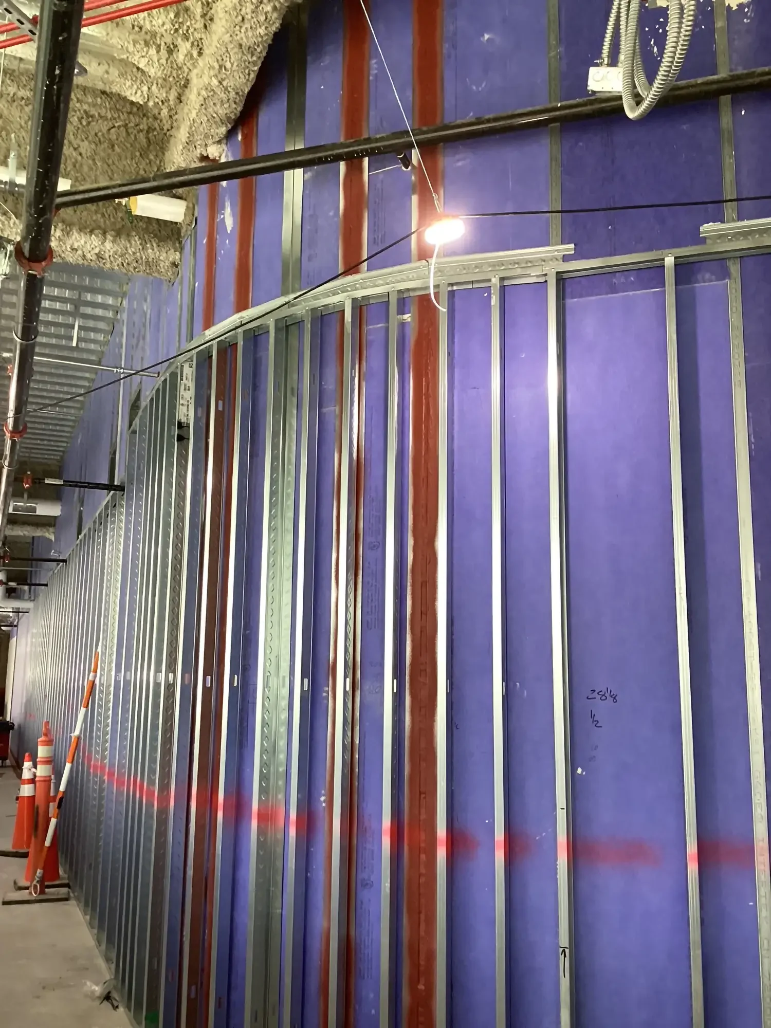 Commercial construction interior with purple insulated wall panels, metal framing studs, exposed pipes, electrical conduits, and temporary work lights.