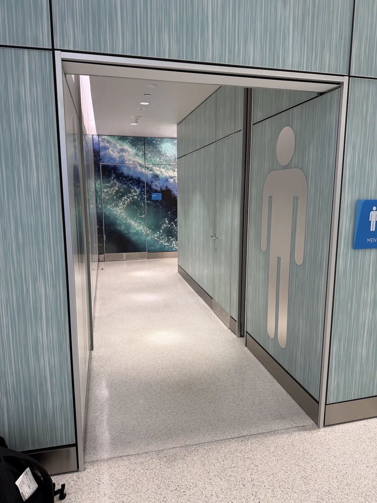 Corridor leading to a men's restroom at an airport, with teal-colored wall paneling, a large stylized man icon on the door, and a blue sign indicating men's restroom