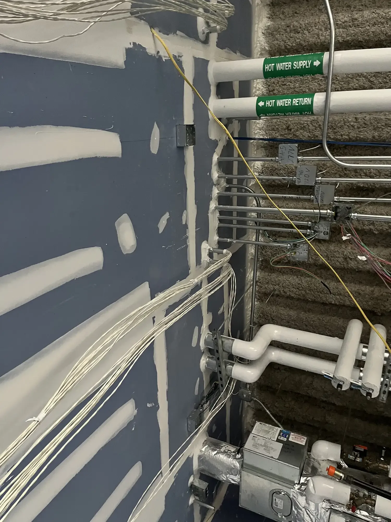 A plumbing and electrical system inside a construction wall. White pipes with green labels for hot water supply and return, wiring bundles, electrical boxes, and other components on a dark blue wall.
