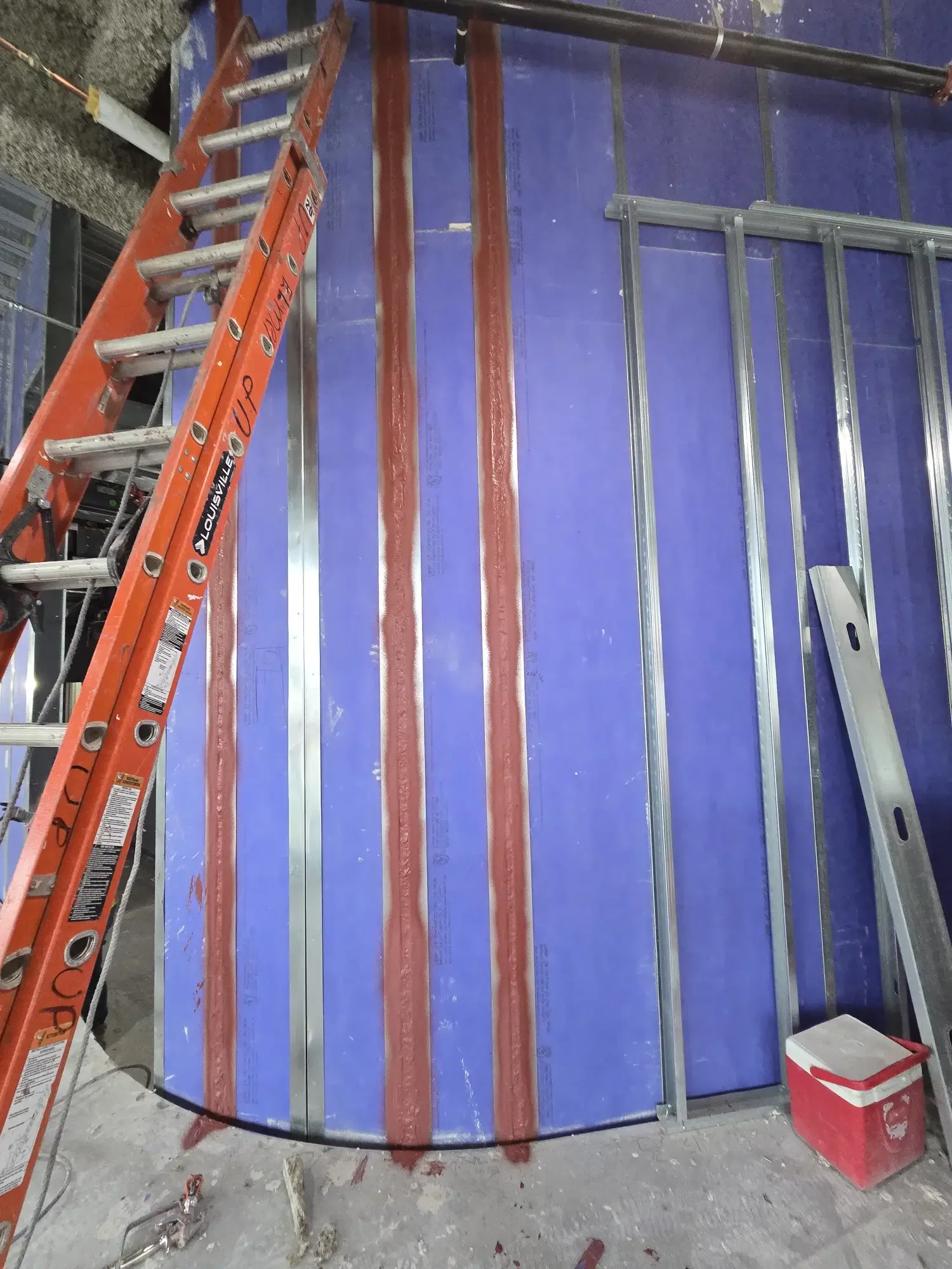 Construction site with an orange ladder on the left, a large blue wall or panel, and a red and white cooler box on the bottom right. Silver metal framing and various tools and materials on the floor.