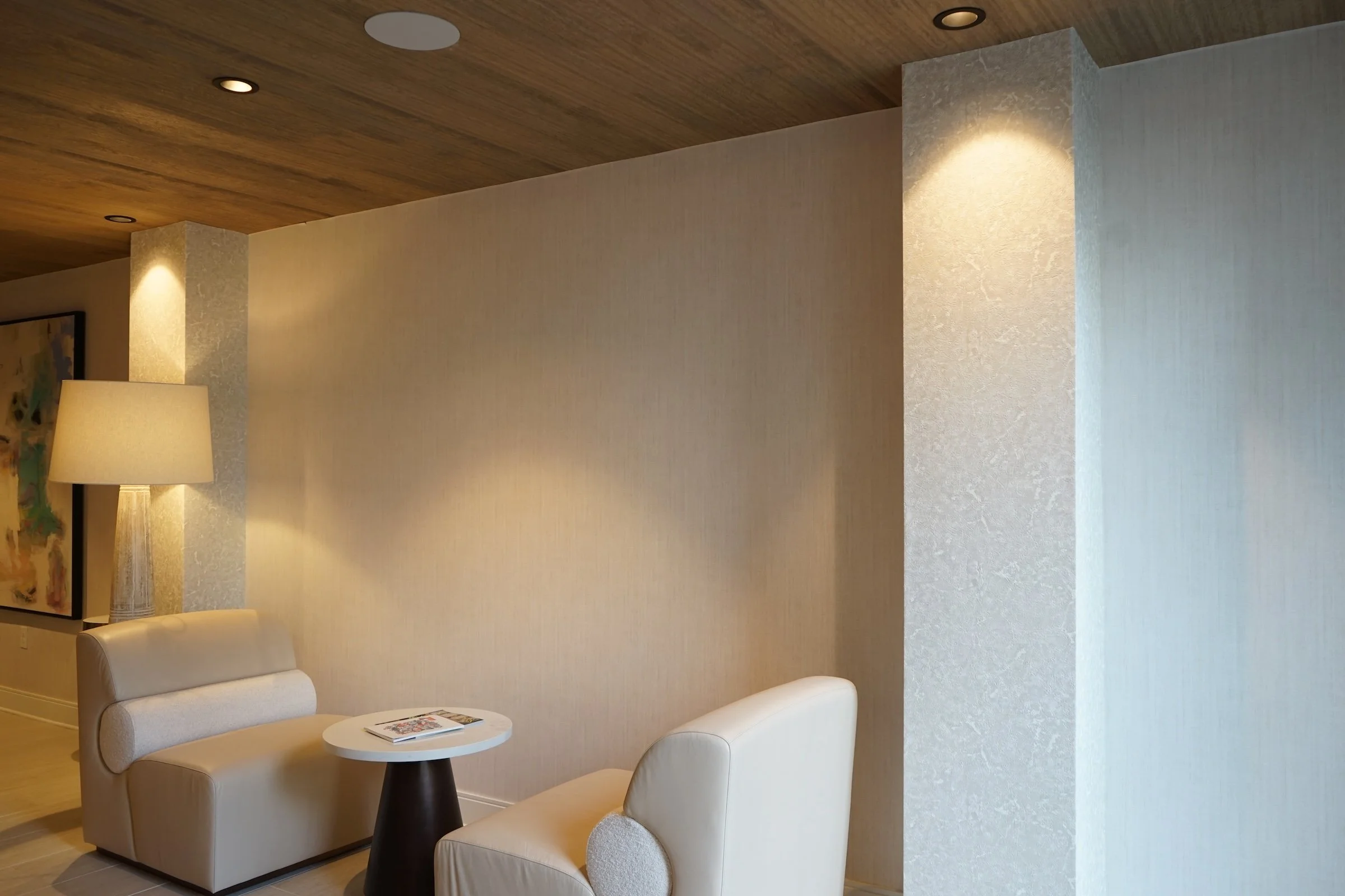 Interior living space featuring cream-colored armchairs, a small round table with magazines, a lamp, a textured beige wall, and a wooden ceiling with recessed lighting.
