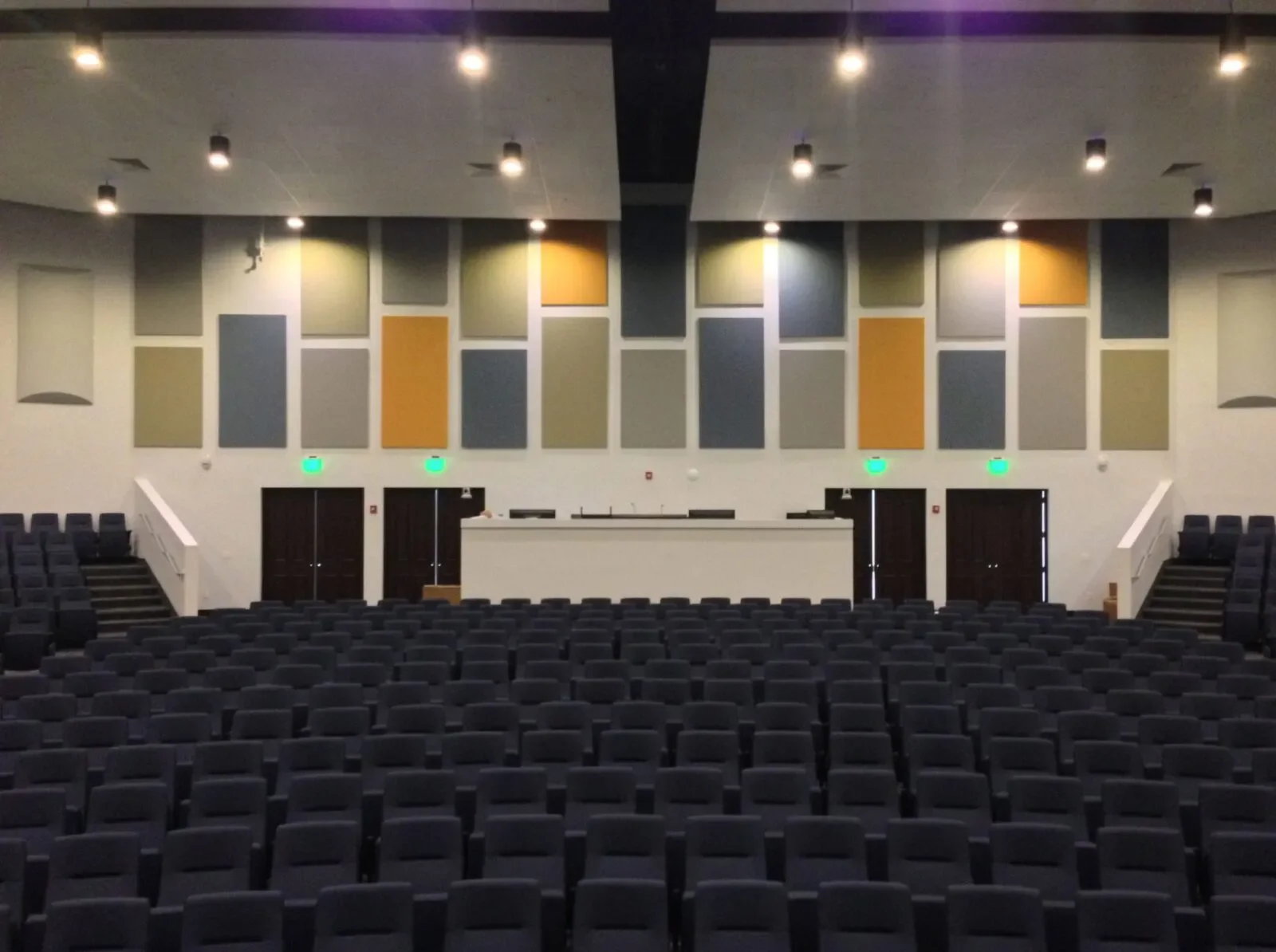 An empty auditorium with rows of black chairs, a stage with a white podium, and a colorful geometric wall design behind the stage.