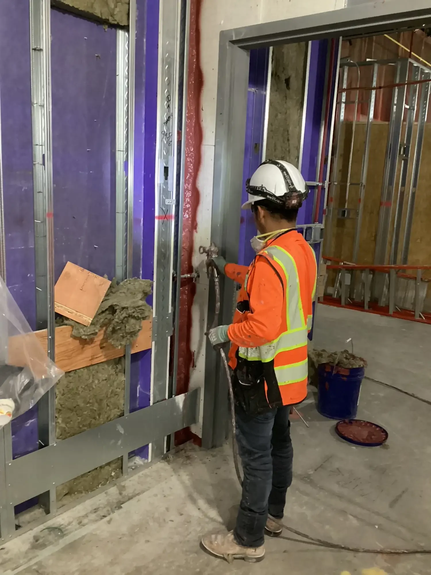 A construction worker wearing a white helmet, orange safety vest, and glasses, spray painting the metal frame of a wall. The setting appears to be indoors at a construction site with exposed framing and insulation.