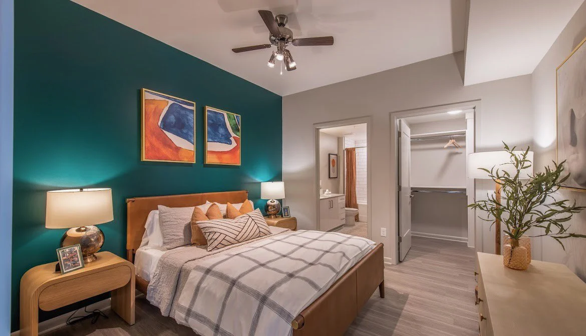 A modern bedroom with a teal accent wall, a wooden bed, four decorative pillows, two bedside lamps, three paintings above the bed, and a doorway leading to a walk-in closet and bathroom. There is a dresser with a plant on it on the right.