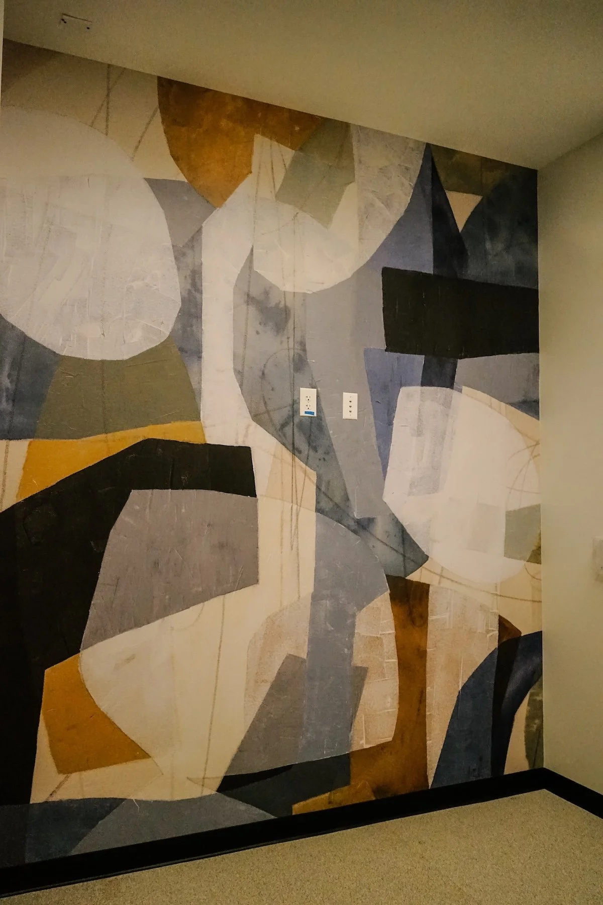 Abstract geometric mural in shades of gray, white, black, gold, and beige covers a wall, with two electrical outlets visible in the center.