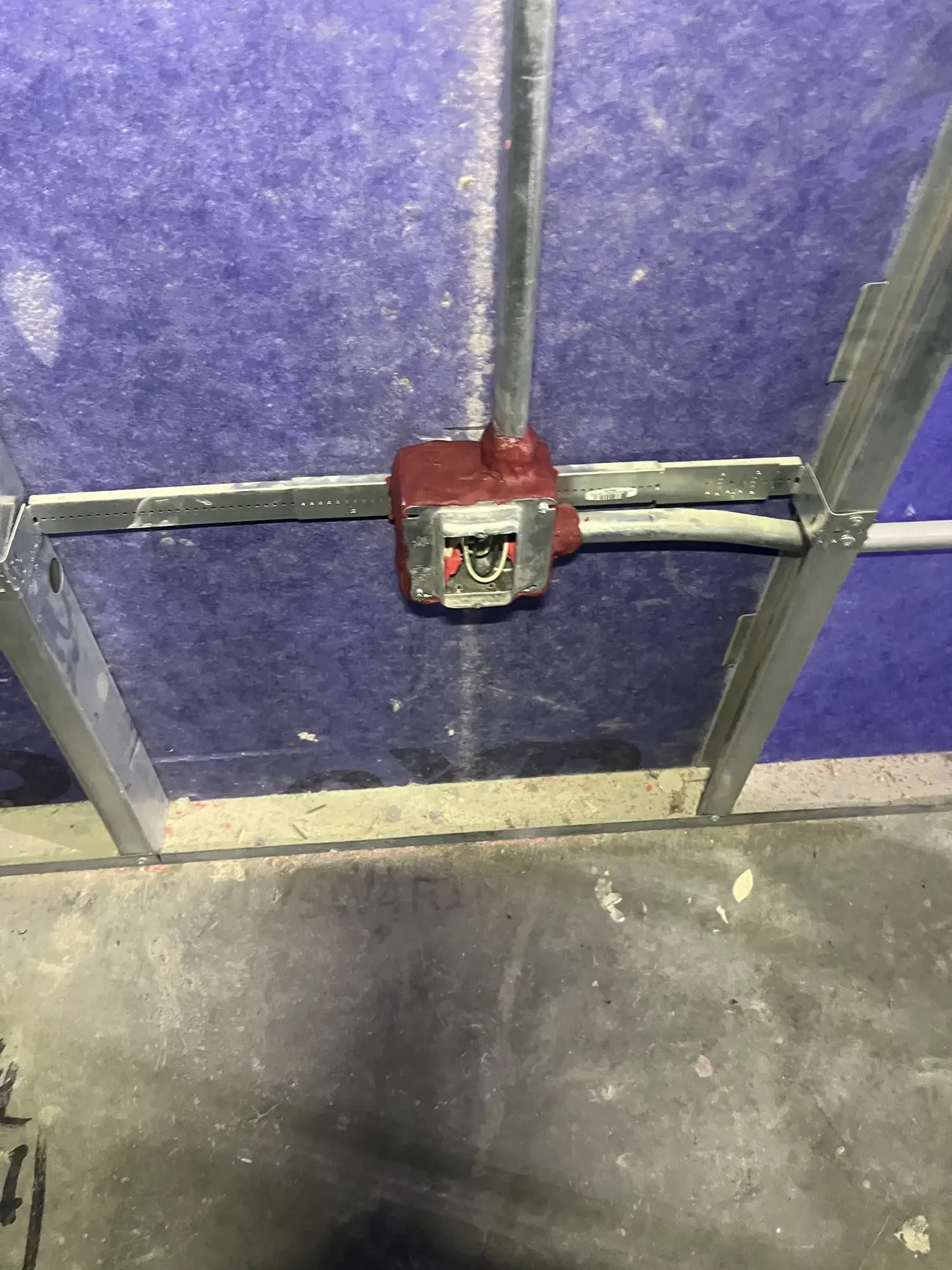 The image shows an unfinished metal door frame with a mounted electrical box at the bottom center. The background is purple, textured, and appears to be a partition wall, with a concrete floor below.