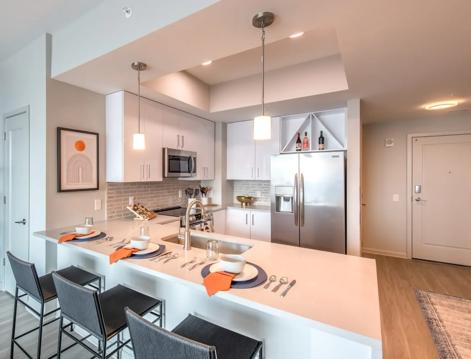 Modern kitchen with white cabinets, stainless steel refrigerator, microwave, and a kitchen island set for four with orange napkins, black placemats, bowls, and utensils. Pendant lights hang above the island, and a framed abstract art piece decorates 
