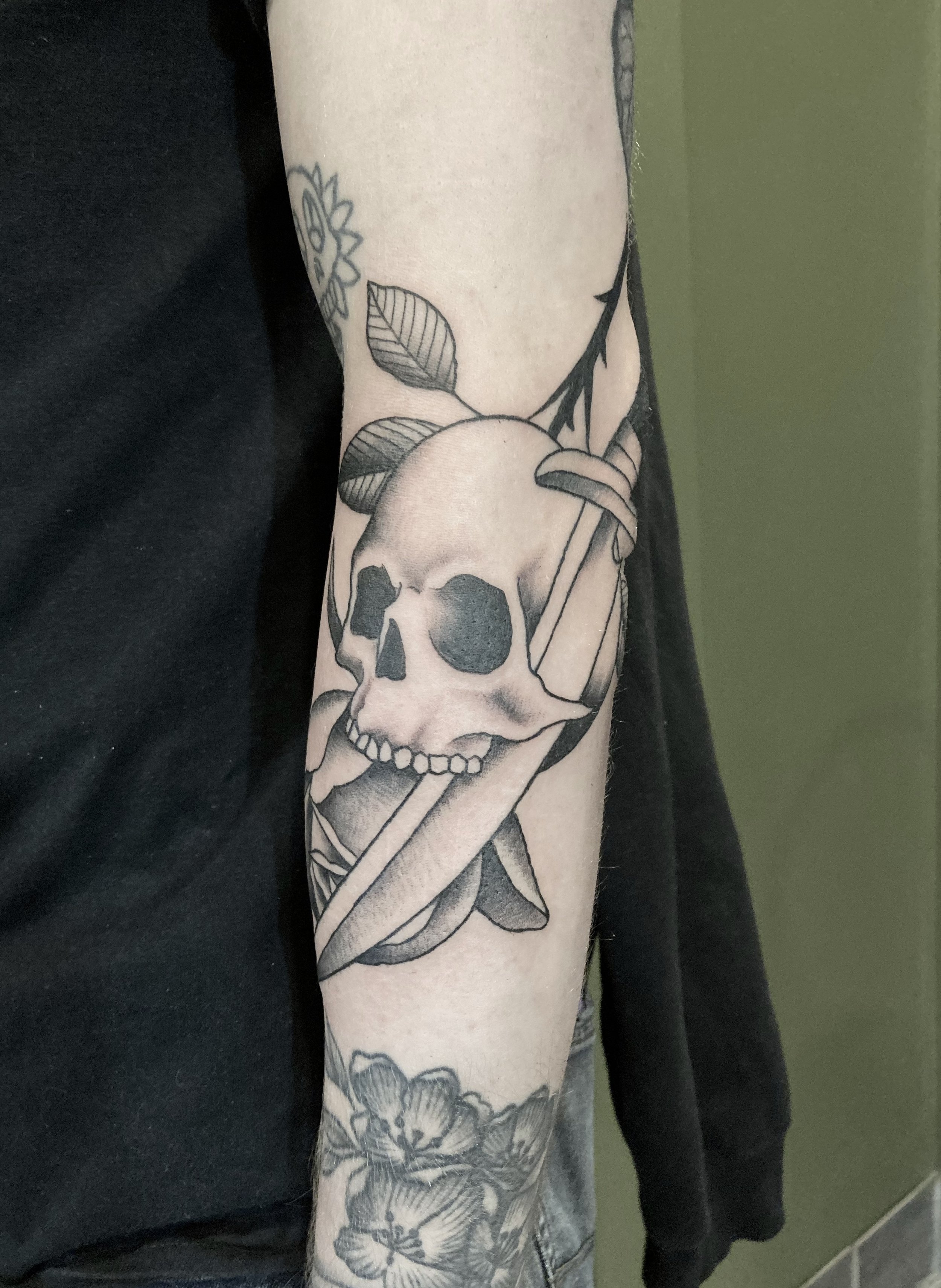 Tattoo of a skull with leaves and a dagger on a person's arm.