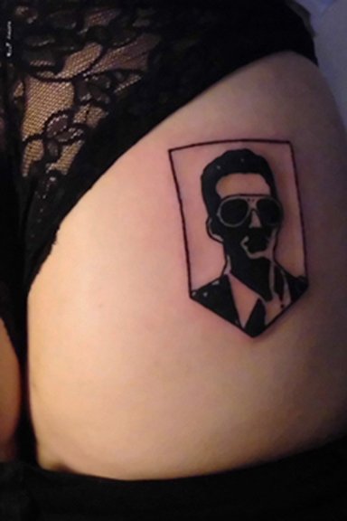 Tattoo of a silhouette of a person with glasses, wearing a suit, inside a shield-shaped border on skin.