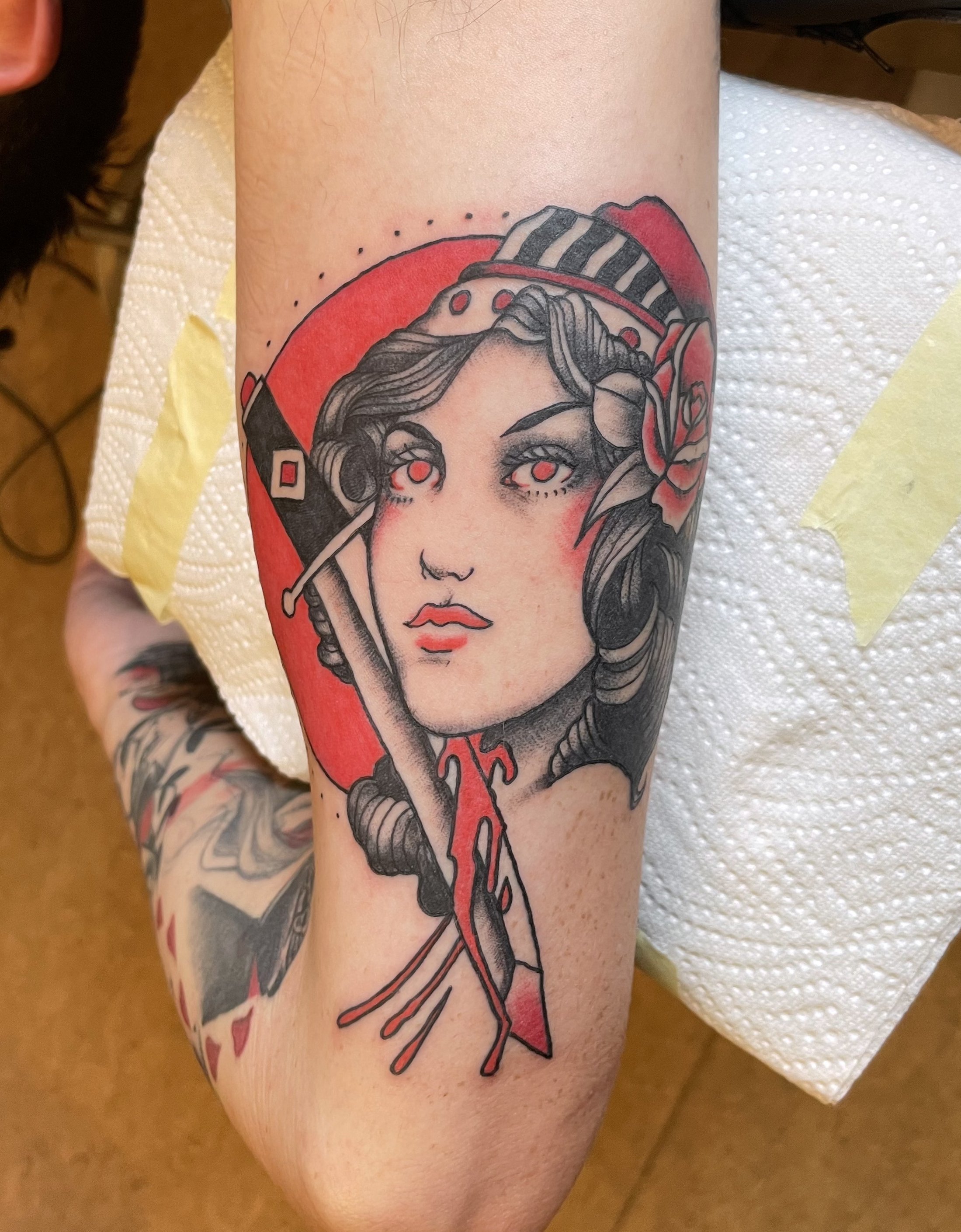 Tattoo of a woman with vintage features, holding a sword, with a red circle background, shaded in black and gray tones with red accents.