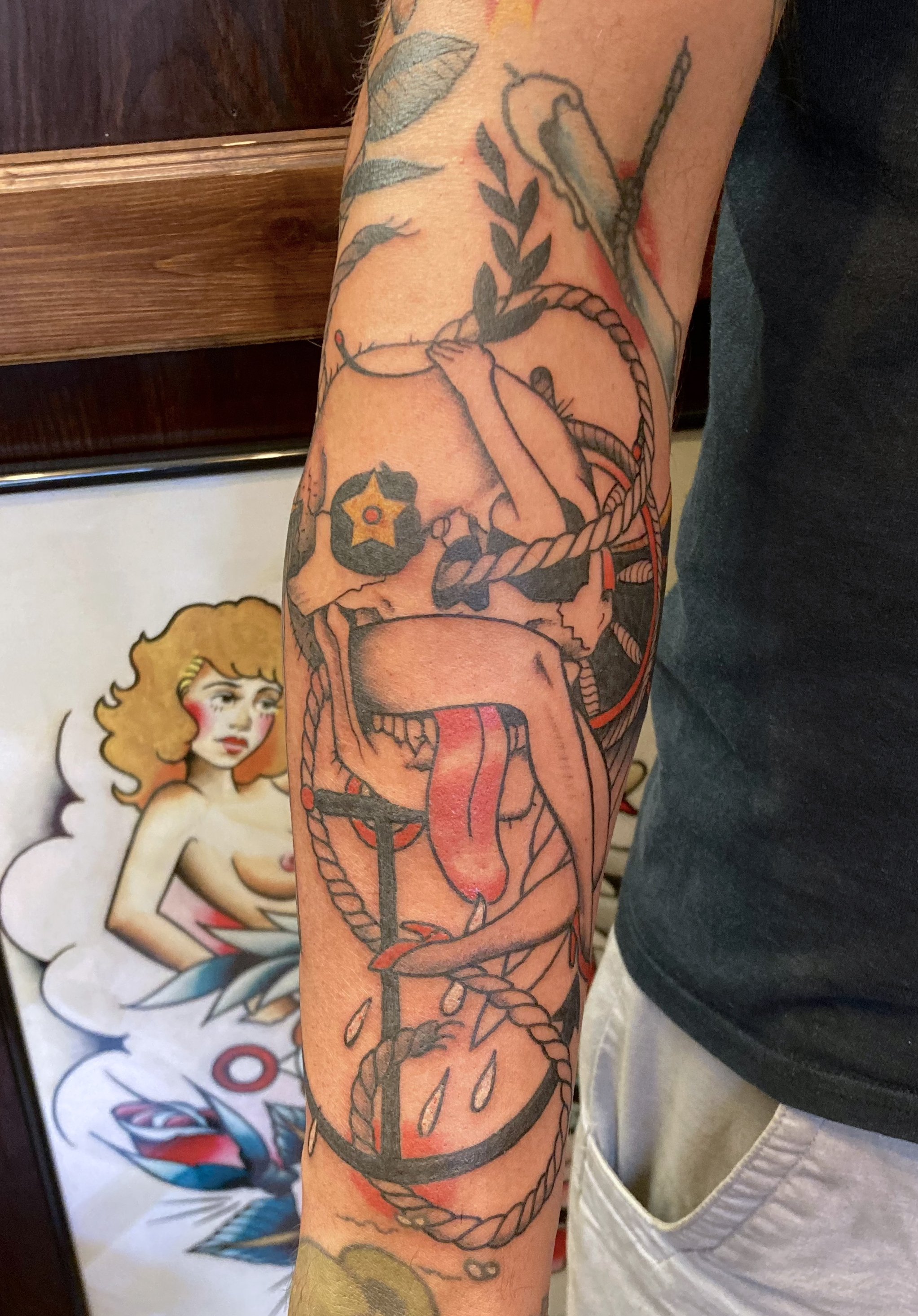 Tattoo of a nude woman with long hair and a star on her breast, holding a rope wrapped around herself, with a red tongue sticking out, on a person's arm.