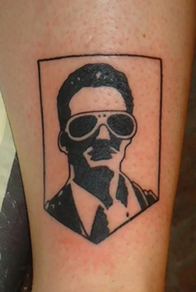 Black and gray tattoo of a man with sunglasses, and styled hair within a rectangular frame on skin.