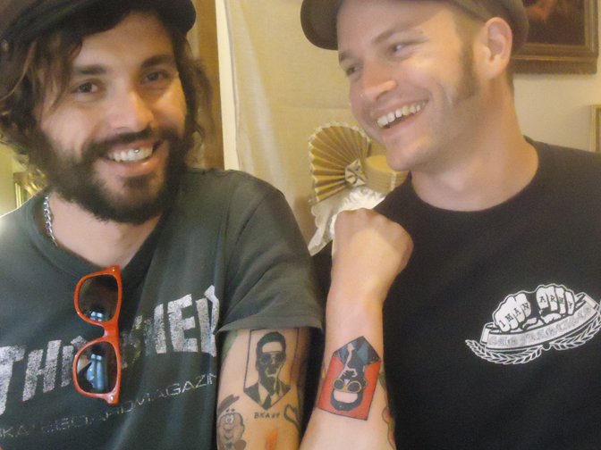 Two smiling men with tattoos, wearing black shirts and hats, sitting indoors with a decorative fan in the background.