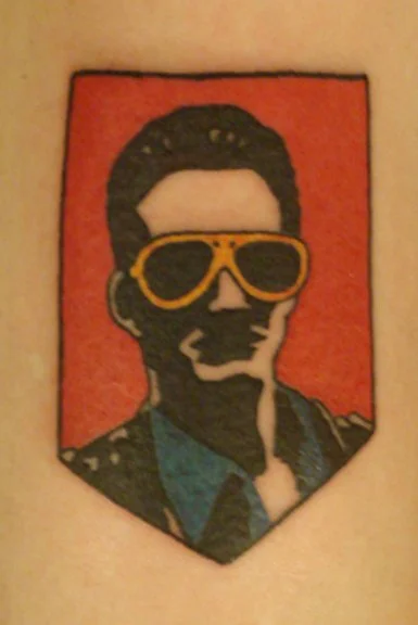 A tattoo of a man wearing sunglasses on the upper arm. The background of the tattoo is red, and the man is depicted with dark hair, and a half-smile.