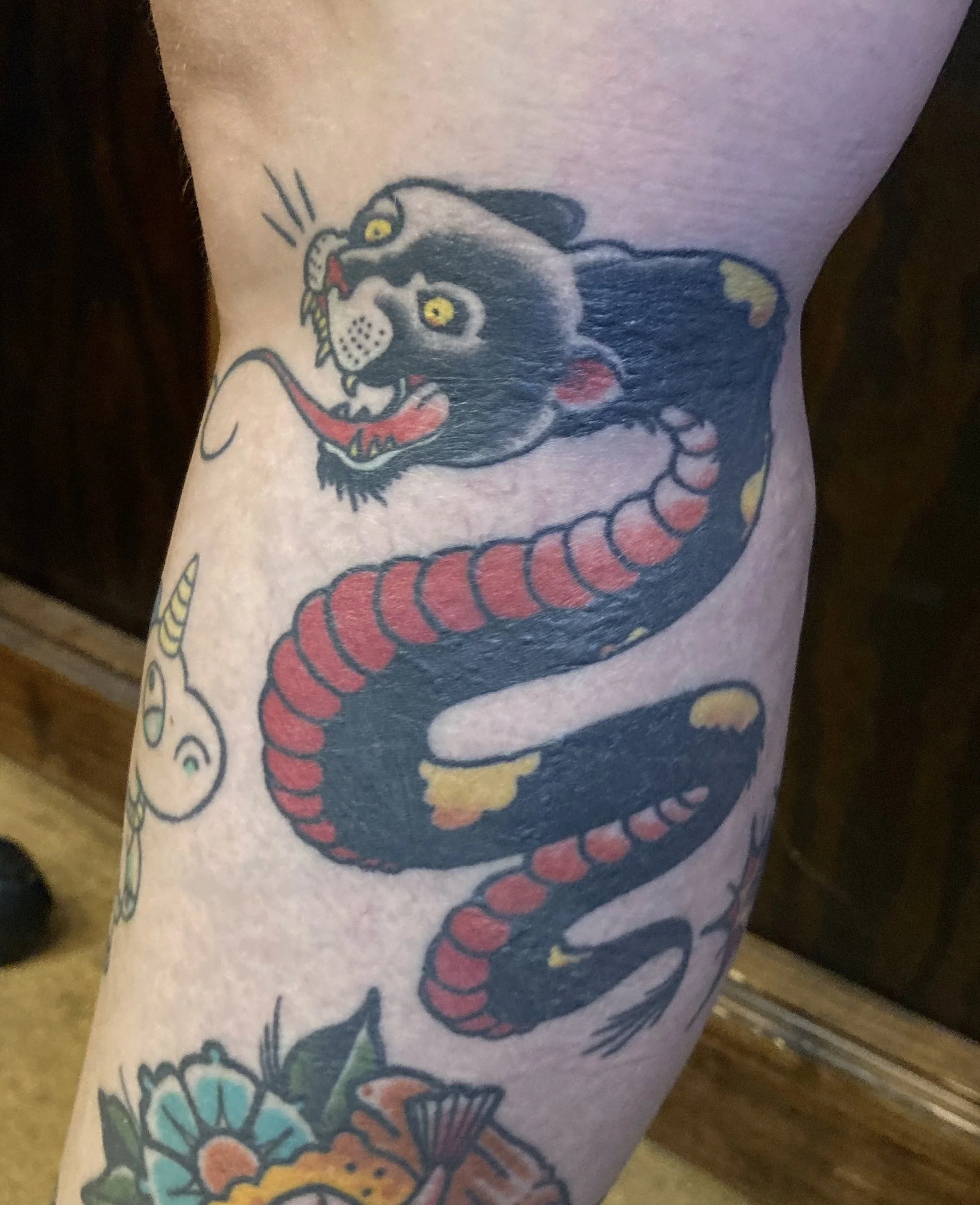 Tattoo of a black panther with yellow eyes, open mouth showing sharp teeth and a long red tongue, on a person's leg.