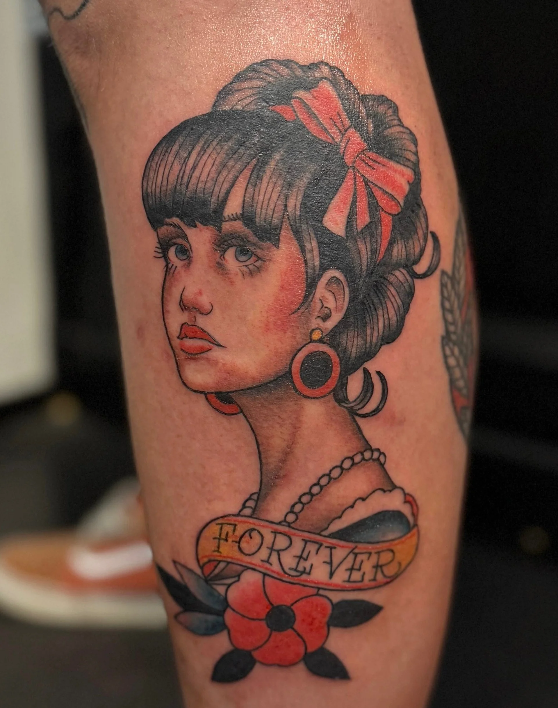 Tattoo of a woman with dark hair, red bow, earrings, and necklace, with a banner reading 'FOREVER' and a red flower with black leaves.