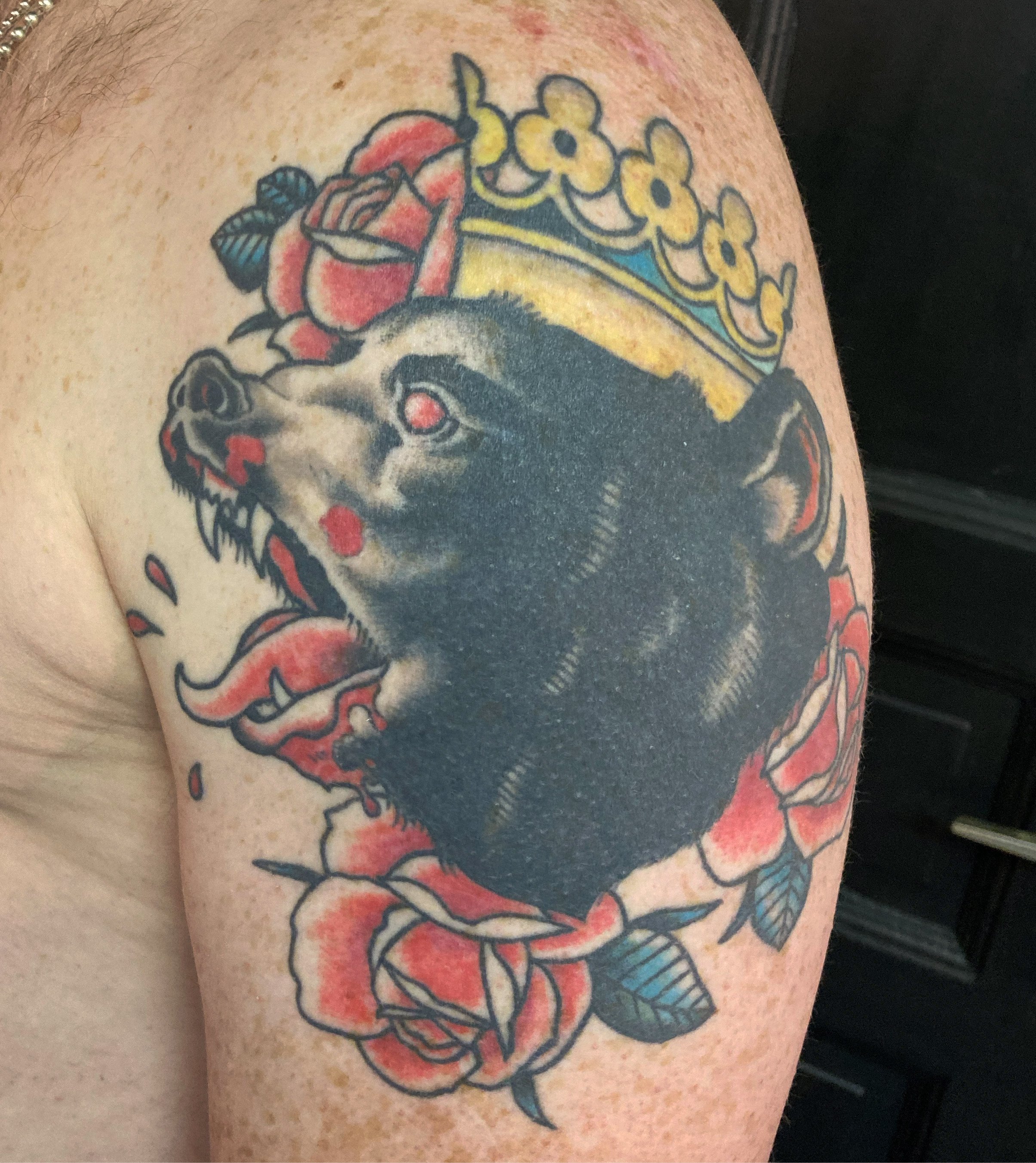 Tattoo of a bear wearing a yellow crown with blue details, surrounded by red roses on the upper arm.