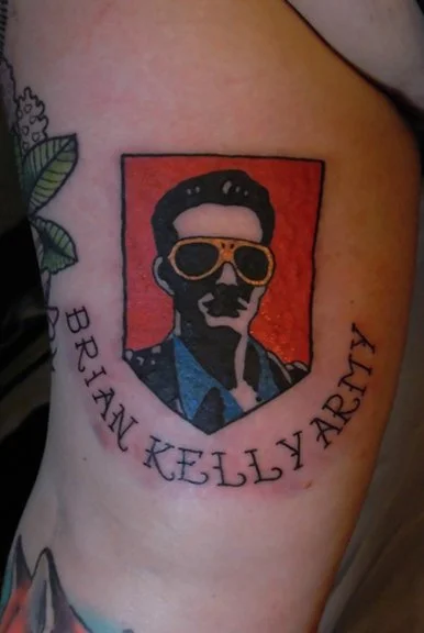 Tattoo of a man with dark hair, sunglasses, wearing a blue shirt and black jacket, inside a shield-shaped design with the text "BRIAN KELLY ARMY" around it.