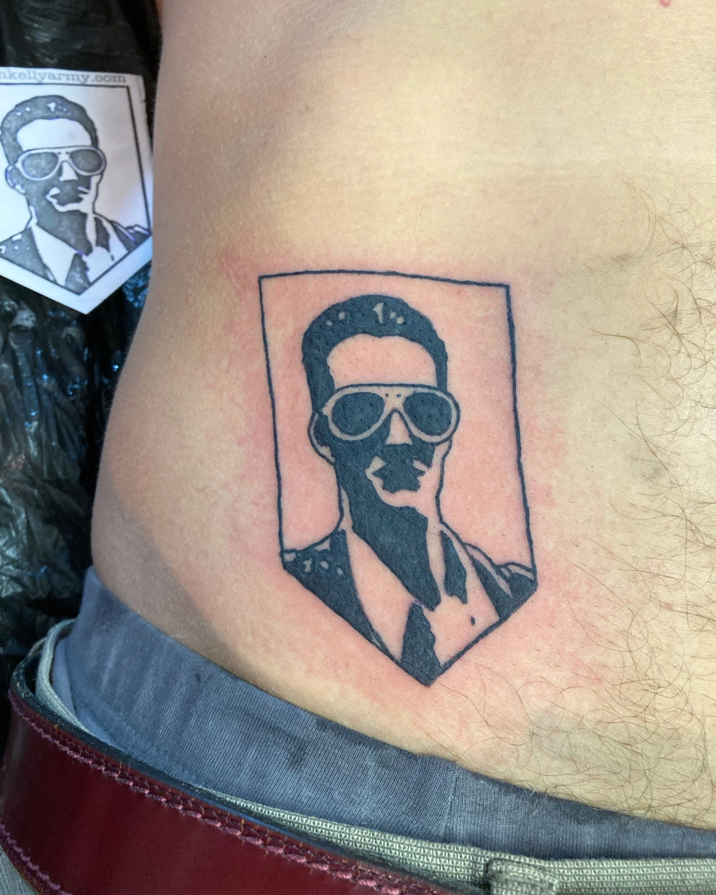 Tattoo of a stylized portrait of a man with glasses, depicted in black ink, enclosed in a rectangular frame on a person's side.