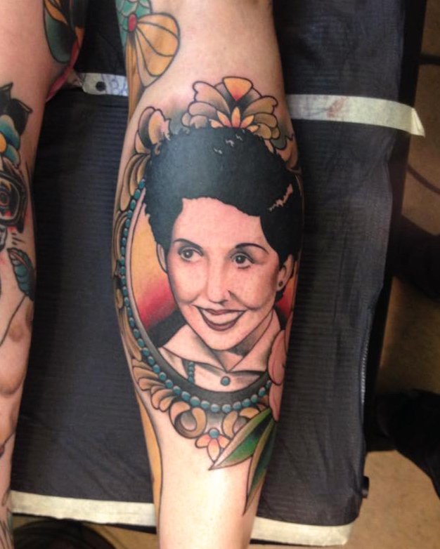 A tattoo of a woman's face with short dark hair and a playful expression, surrounded by floral decorations and a beaded necklace tattoo. The tattoo covers most of the person's lower leg.