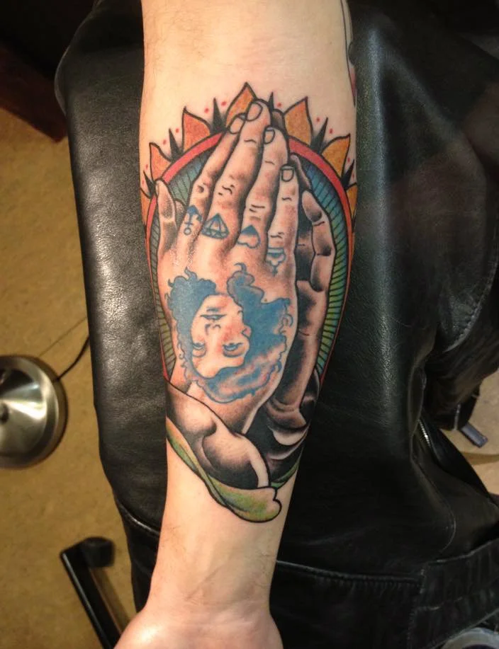 Tattoo of a woman with curly hair, partially covered by a large hand with several rings, framed by a colorful sunburst design.