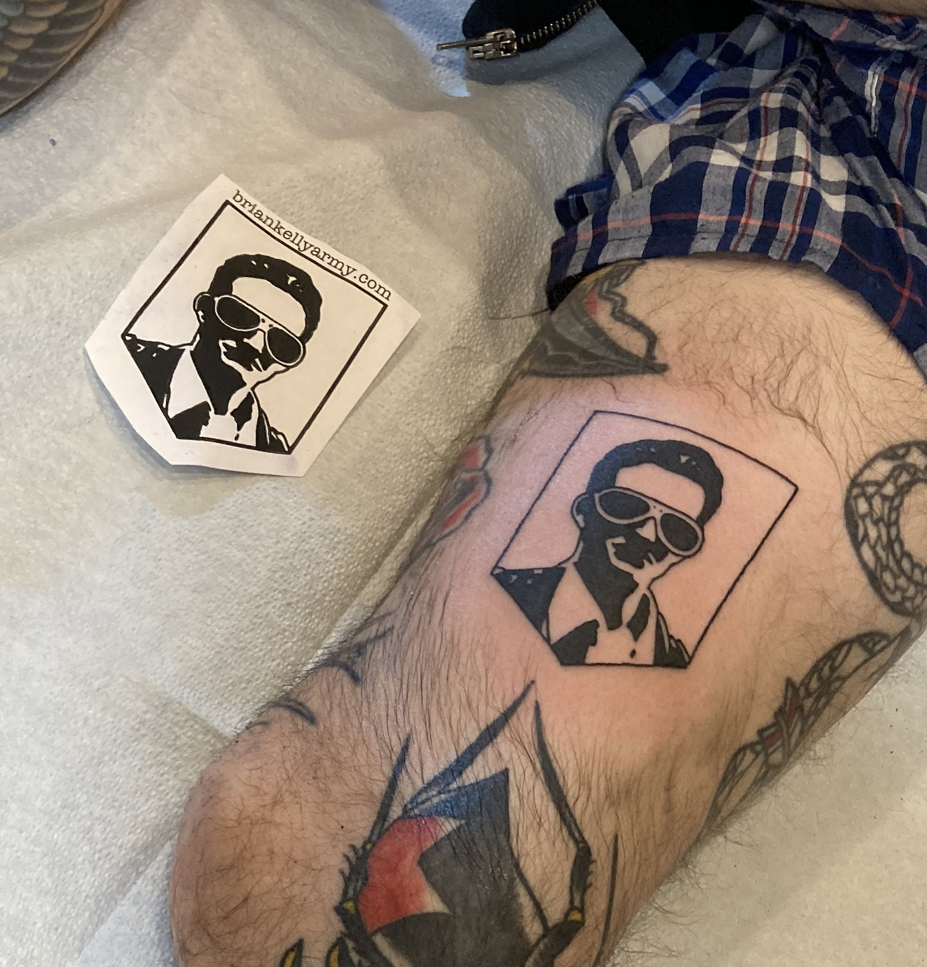 A tattoo of a black and white portrait of a man wearing sunglasses, within a rectangular frame on a person's thigh, with a sticker of the same design nearby.