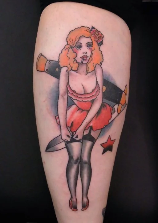 Tattoo of a woman with red hair, pink flower in hair, wearing a pink top and red skirt, standing with legs close together, holding a large knife pointing downward, with star and other decorative elements around.