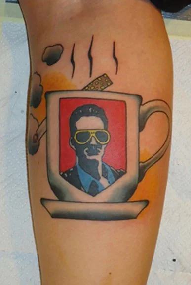 Tattoo of a coffee cup with a picture of a man wearing sunglasses inside, with steam rising from the cup.
