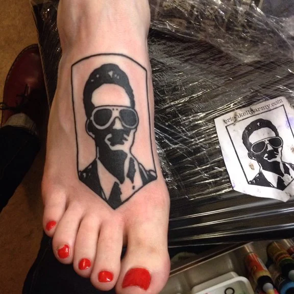 Tattoo of a person wearing sunglasses and a suit, with a hairstyle reminiscent of Elvis Presley, on a person's foot.