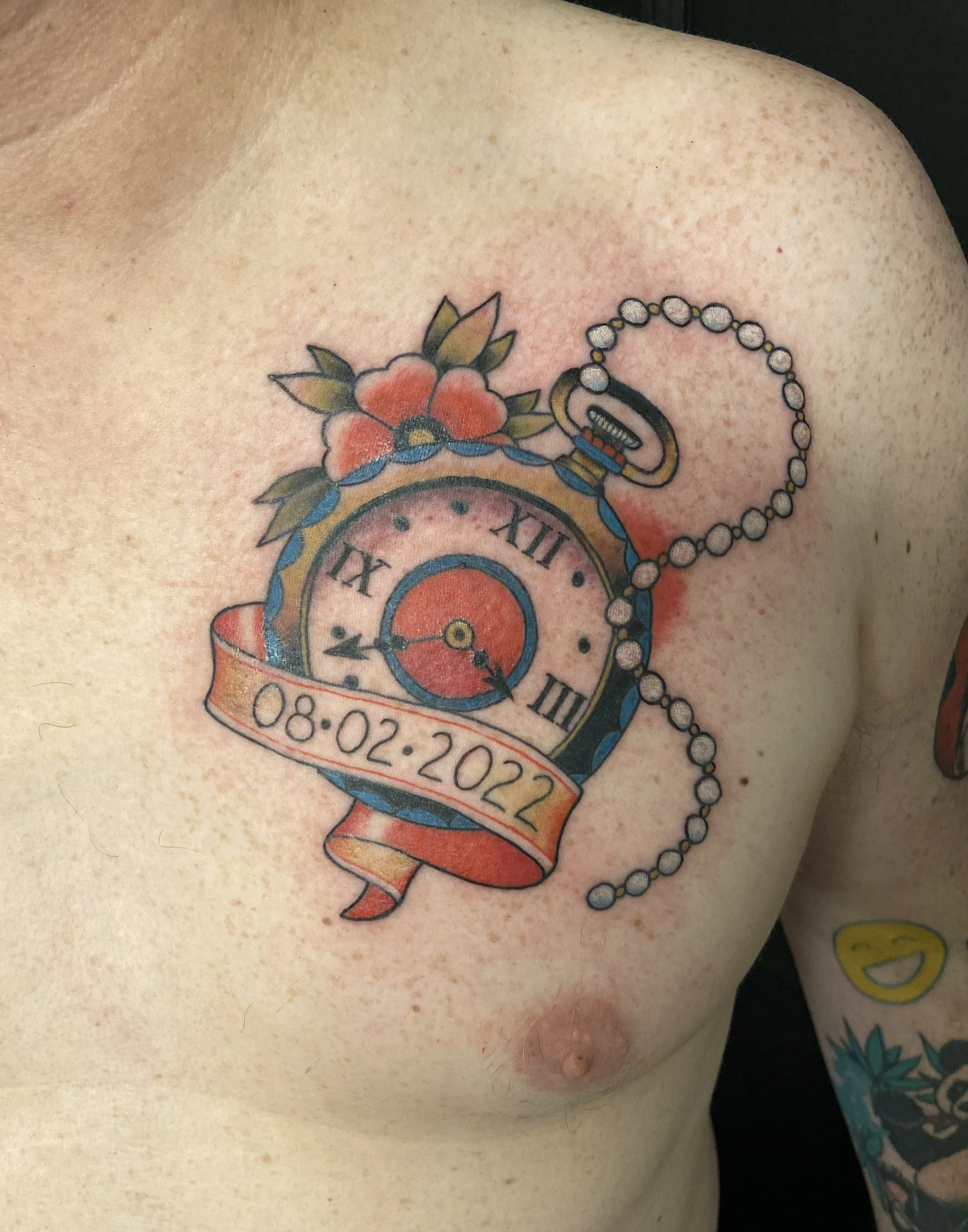 Tattoo of a vintage pocket watch with Roman numerals, a chain, and a ribbon displaying the date 08-02-2022 on a person's chest, with floral accents.