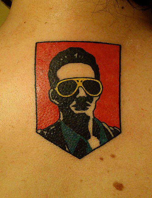 Tattoo of a man with sunglasses, and styled hair on an upper back, against a red background.