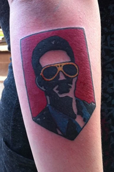 Tattoo of a stylized portrait of a man wearing sunglasses with a red background.