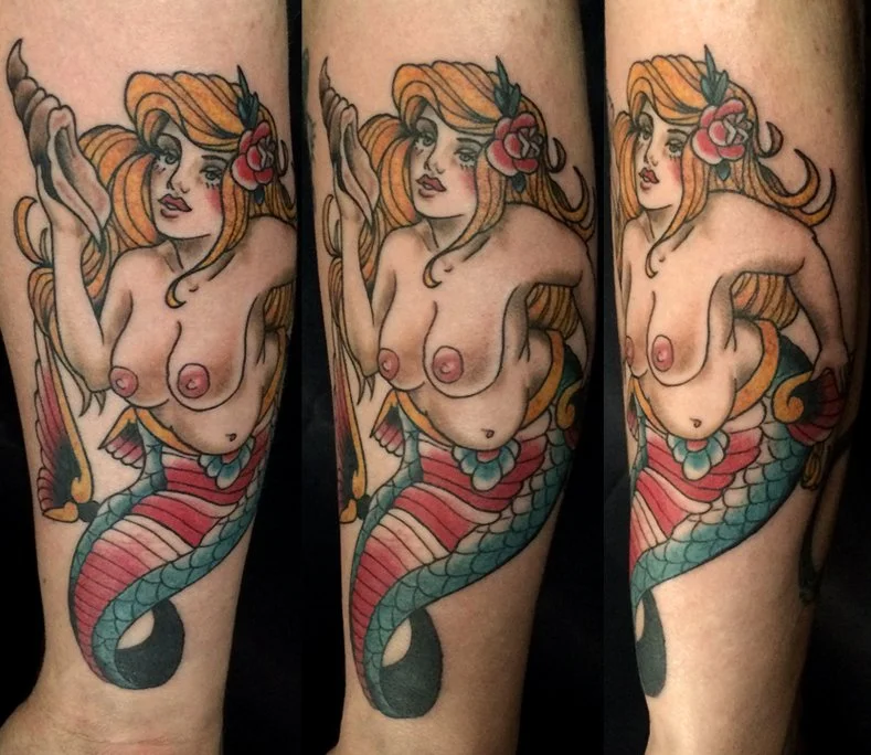 Tattoo of a mermaid with long, flowing orange hair, pink flowers, and a colorful tail, depicted in a traditional tattoo style.