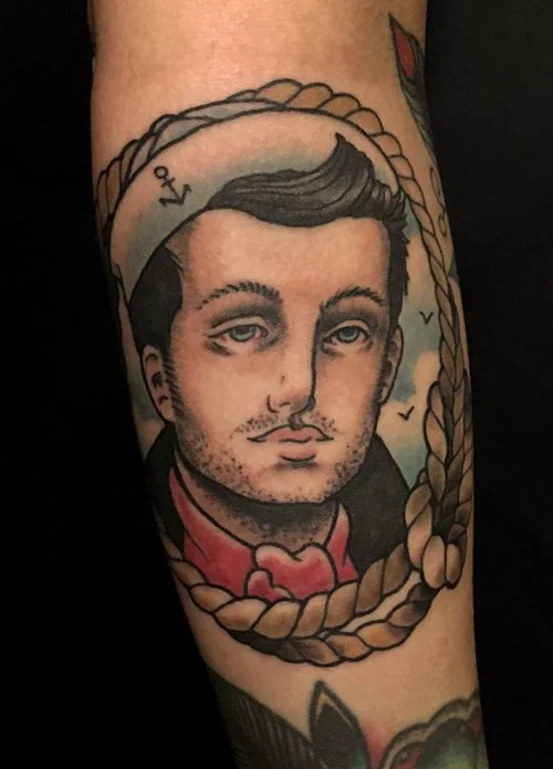 Tattoo of a man's portrait with blue eyes and styled hair, surrounded by a rope frame, on a person's arm.