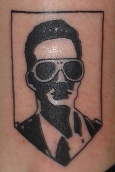 Black tattoo of a person wearing sunglasses and a leather jacket, within a rectangular border.